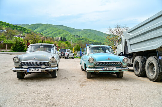 Two historic GAZ-21 Volga classic cars from the 1950s, in light gray and light blue, stand side by side in a parking lot in front of green hills near Shignaghi; Soviet classic cars, Georgia