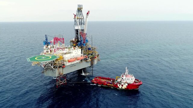 Wide aerial shot of an offshore oil rig platform in the ocean with a red supply ship anchored alongside for logistics and maintenance operations.