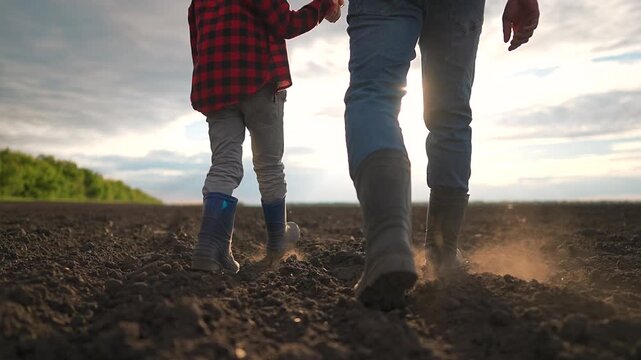 Father walks with child through field under dramatic sunset sky. Both wear matching shirts and caps. Scene shows close father and child connection while walking through quiet nature together.