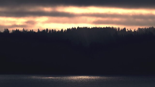 4K Sun Rays Shining Through Silhouetted Pine Tree Forest Reflected in Still Lake at Sunrise