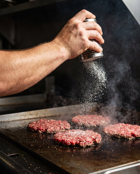 Chef Seasoning Fresh Raw Burger Patties on Sizzling Griddle with Salt Shaker