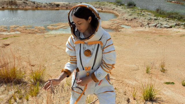 Astrobiologist in a spacesuit examining dry plants on the shore of an alien lake