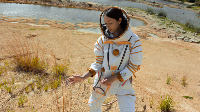 Alien flora discovery as a spacesuited woman reaches toward grass by a quiet lake