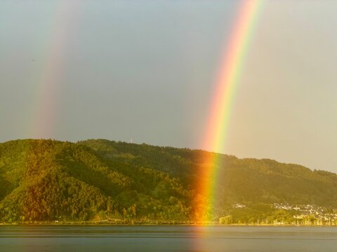 Vibrant double rainbow over a calm lake and green hills during golden hour
