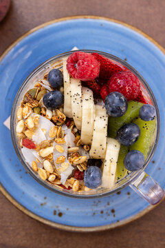 layered yogurt bowl served in clear cup with bananas, kiwis, raspberries, blueberries, chia seeds, and granola on a blue ceramic plate