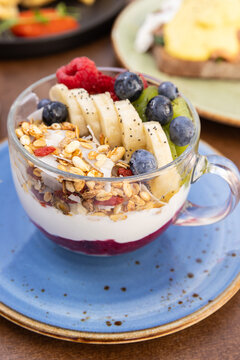 layered yogurt bowl served in clear cup with bananas, kiwis, raspberries, blueberries, chia seeds, and granola on a blue ceramic plate