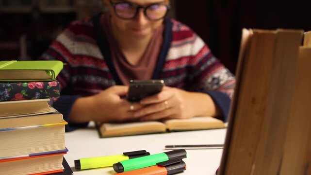 Student studying at desk with books and phone. A focused female student studying at a desk in the evening, surrounded by books and stationery, checking her smartphone while working on an assignment