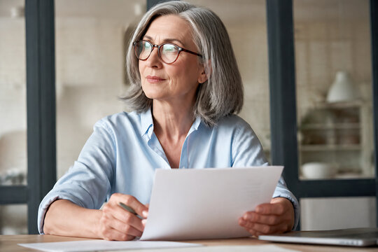 An older woman wearing glasses is sitting at a table, looking thoughtfully at some documents.
