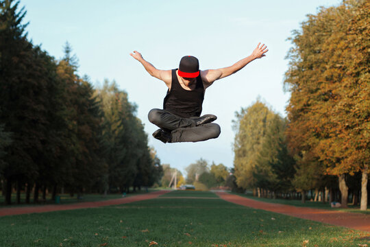 Athletic male dancer frozen in mid-air during a high jump with crossed legs in a park alley, demonstrating impressive agility, strength, and street style in warm sunlight.