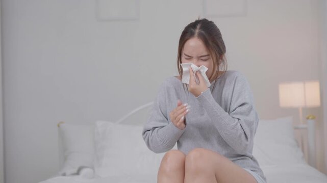 	
Cold, sickness and flu symptoms concept. Young Asian woman sneezes into a tissue while sitting on a bed at home, feeling unwell. Cold, sneezing, sickness, flu, healthcare