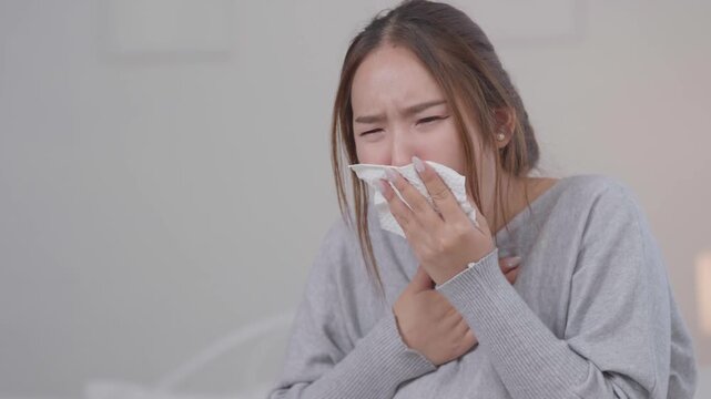 	
Cold, sickness and flu symptoms concept. Young Asian woman sneezes into a tissue while sitting on a bed at home, feeling unwell. Cold, sneezing, sickness, flu, healthcare