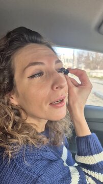Young woman applying mascara to eyelashes while sitting inside a car during the day. Close-up static shot.