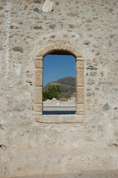 A view of a rocky hill and clear blue sky framed by an ancient stone wall window opening of a historic fortress in Kythira.