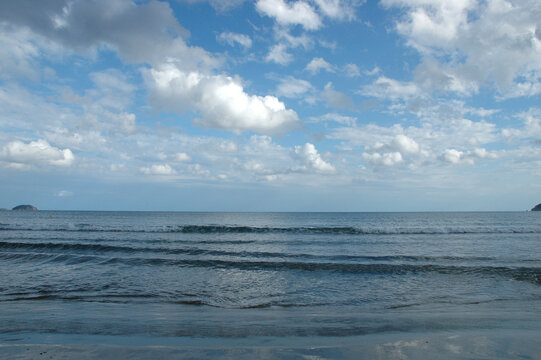 Small ocean waves rolling towards the shore under a bright blue sky filled with white fluffy clouds over the horizon in Greece.