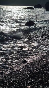 Ocean waves crash against a dark pebble beach at sunset, shimmering under sunlight. Static wide shot captures the rhythmic ebb and flow movement.