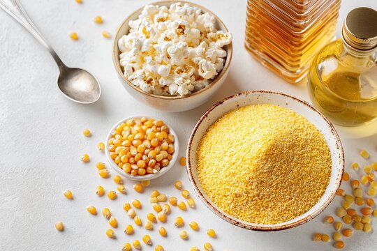 Raw corn kernels, dried popcorn in bowl, yellow cornmeal, vegetable oil bottle, metal spoon on white background