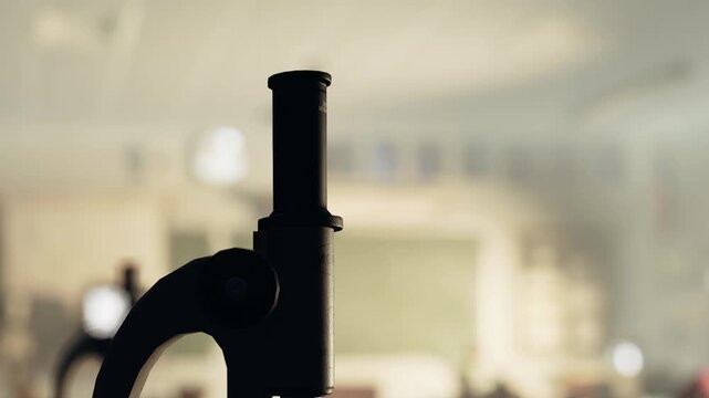 A machine part stands prominently in a workshop filled with tools and equipment. Light shines through a window, illuminating the workspace. Workers may be present but are not seen.