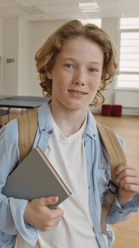 Vertical portrait of Caucasian teenager boy holding book and backpack, smiling at camera. Clip is ideal for school enrollment promotions and educational marketing campaigns