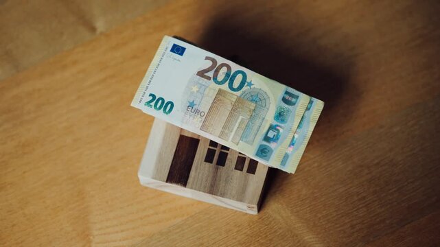 A close-up of a stack of euro bills being placed on a small wooden house; buying real estate and a mortgage loan
