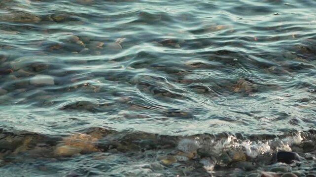Pebble shore ripples under clear water waves during daytime. Static close-up shot.