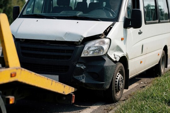 White minibus shows front crash damage on road. Headlight, bumper smashed bending metal hood with visible bumper collision. Tow truck lifts vehicle for insurance claim repair measure. Hands-free