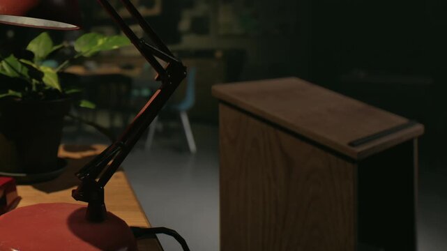 A lamp with a red base sits on a wooden desk. A green plant appears beside it. In the background, there is a blue chair and blurred items on shelves. The room is dimly lit.