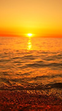 Golden light glimmers over ocean waves rolling onto a pebble shoreline during an evening dusk. Static vertical close-up shot.