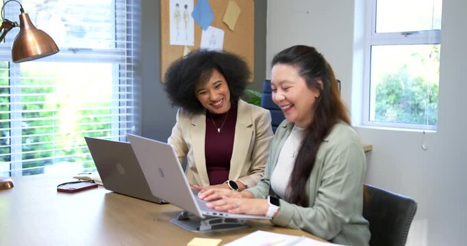Diverse female coworkers reviewing project draft, right woman typing on raised laptop stand at desk