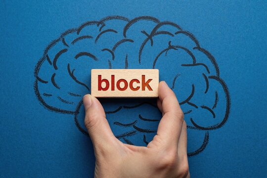Hand places wooden block labeled block onto simplified brain drawing. Symbolizes mental block or creative stagnation hindering thought process and problem solving.