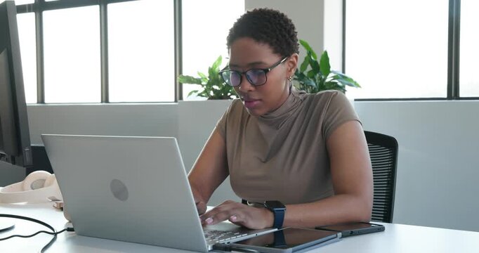 African American adult woman typing on laptop at desk, reading screen, pausing, wearing smartwatch