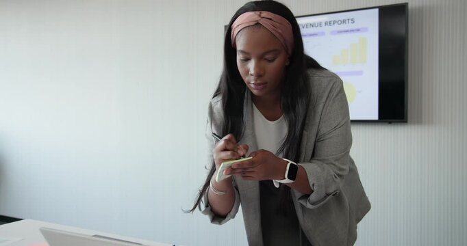 African American woman in blazer in meeting room responding to sales slide, writing sticky notes