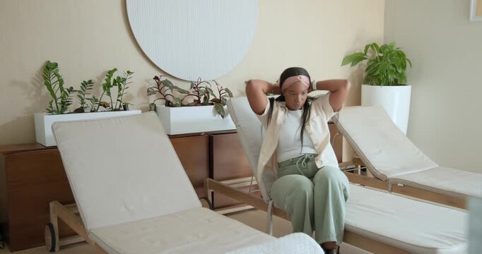 African American woman entering spa room walking to chair sitting fixing headband seeking rest