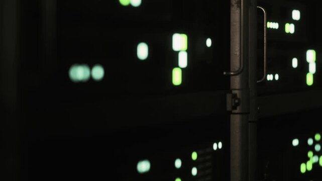 Green lights blink on digital devices in a dark data center. The scene shows multiple rows of machines working. The focus is on the bright spots of light against the dark background.