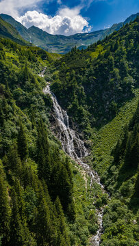 Aerial view of a cascading waterfall flowing through a lush green mountain valley surrounded by dense pine forests near the Transfagarasan in Curtea de Arges, Judetul Arges, Romania.