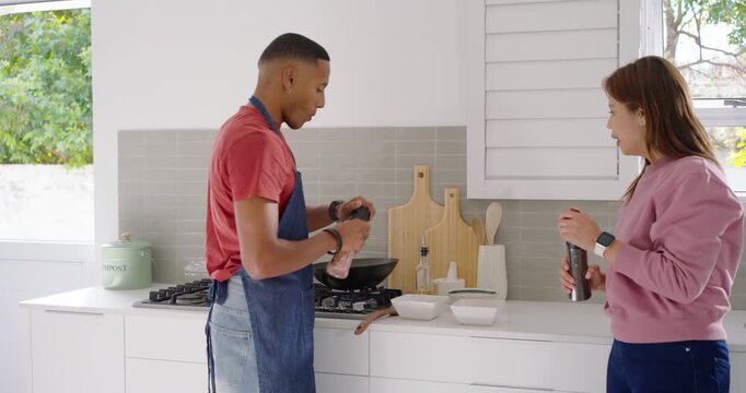 African American couple woman handing grinders while aproned man seasoning wok for meal in kitchen