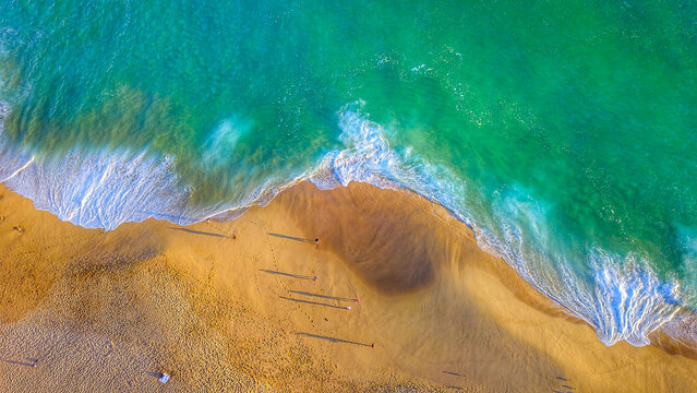 Aerial view of turquoise ocean waves crashing onto the golden sandy beach with long shadows of people walking along the shore in Cabo San Lucas, Baja California Sur, Mexico.