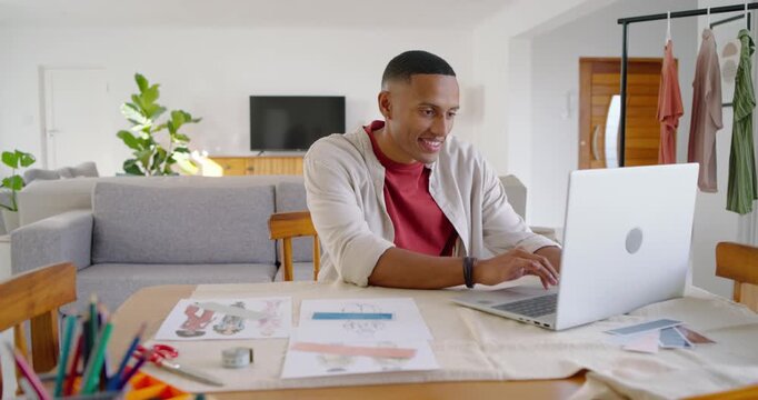 African American man on laptop typing, sorting sketches matching swatch for design at dining table