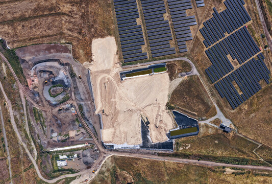 Aerial view of a solar farm with rows of photovoltaic panels next to an active construction site, sandy soil, excavation areas, and retention ponds in Leipzig, Saxony, Germany.