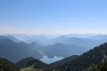Fototapeta premium Atemberaubender Weitblick von einem Berggipfel auf einen tiefblauen, eingebetteten Bergsee, umgeben von dichten Nadelwäldern und gestaffelten Alpenketten im Dunst.