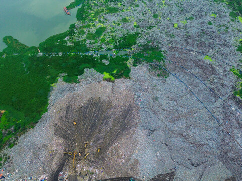 Aerial view of Amin Bazar Waste Disposal Area with heavy machinery working on large piles of waste surrounded by lush green vegetation and water in Bongaon Union, Dhaka Division, Bangladesh.