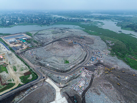 Aerial view of Amin Bazar Waste Disposal Area with heavy machinery operating on large waste piles near the river in Bongaon Union, Dhaka Division, Bangladesh.