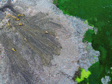 Aerial view of Amin Bazar Waste Disposal Area with heavy machinery and excavators working on piles of waste near lush green vegetation in Bongaon Union, Dhaka Division, Bangladesh.