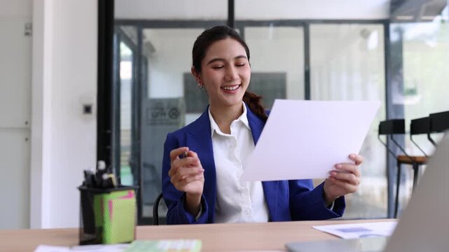 A smiling young Asian businesswoman wearing a blue blazer working in a modern office, happily reading and reviewing a financial report document at her desk with a laptop.