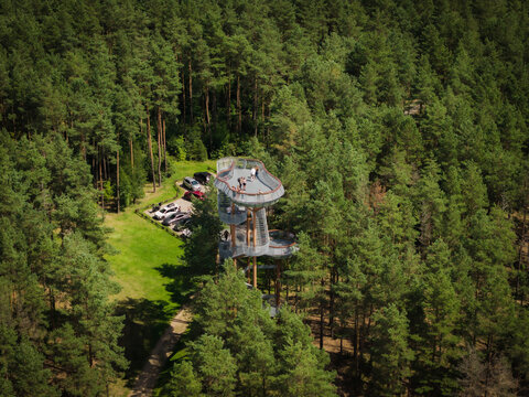 Aerial view of the Merkine observation tower rising above a dense pine forest with a small parking area and visitors on the viewing platform in Merkine, Alytus County, Lithuania.