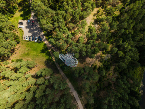 Aerial view of the Merkine observation tower surrounded by a lush pine forest with a nearby parking lot and walking path in Merkine, Alytus County, Lithuania.