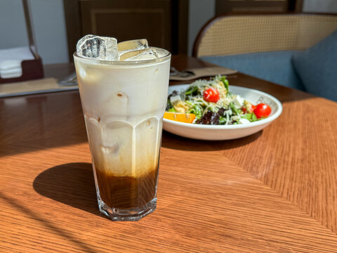 Iced layered protein coffee drink in tall glass on sunlit wooden table with fresh salad in background, soft restaurant interior, casual dining mood, warm daylight, clean food presentation