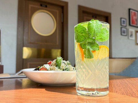Tall sparkling citrus mint drink in clear textured glass on wooden table beside blurred fresh salad inside bright restaurant with warm daylight, inviting refreshing dining atmosphere and elegant