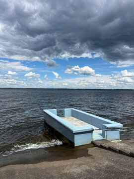 A blue concrete pier on the shore of a wide river or reservoir under a dramatic cloudy sky.
