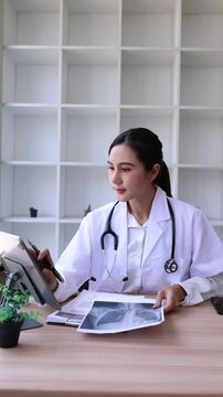 Asian female doctor using digital tablet for online video call consultation with patient and reading medical report in hospital office.