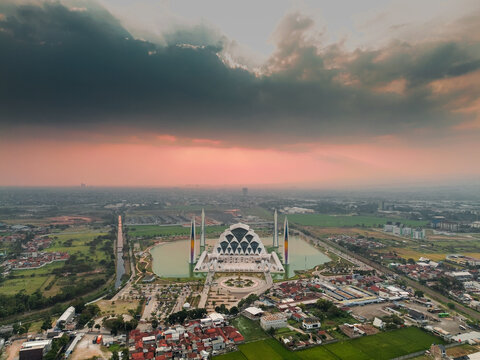 Aerial view of Masjid Raya Al Jabbar surrounded by a reflective pond, lush green fields, and urban rooftops under a dramatic sunset sky in Bandung, Jawa Barat, Indonesia.